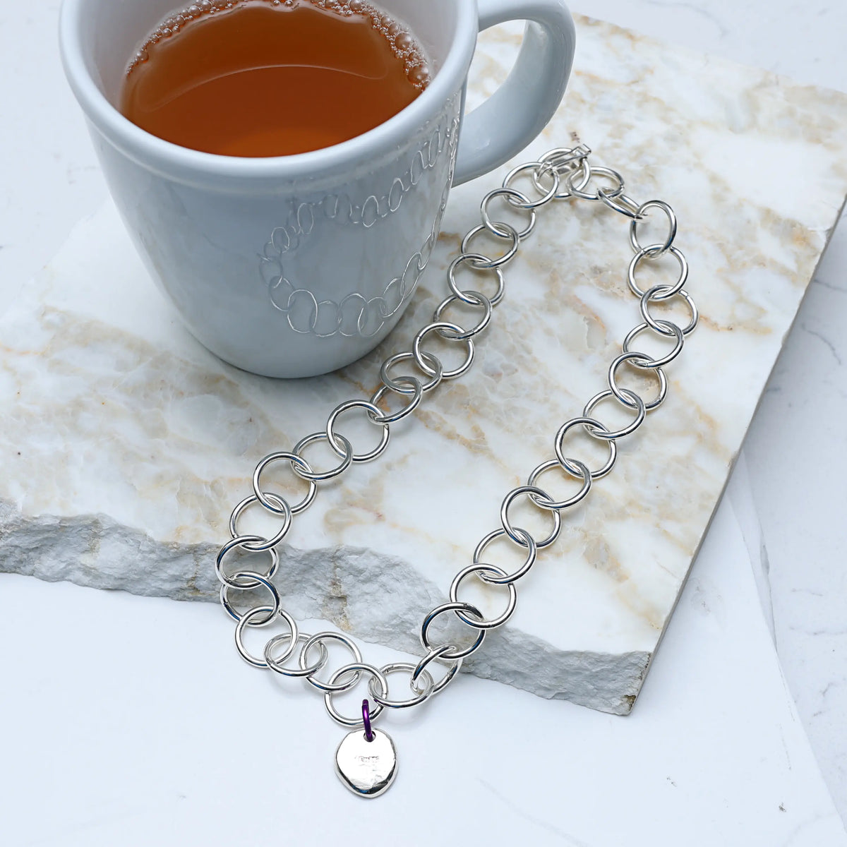 Silver chain necklace with a pendant on a marble surface next to a white mug with a beverage.