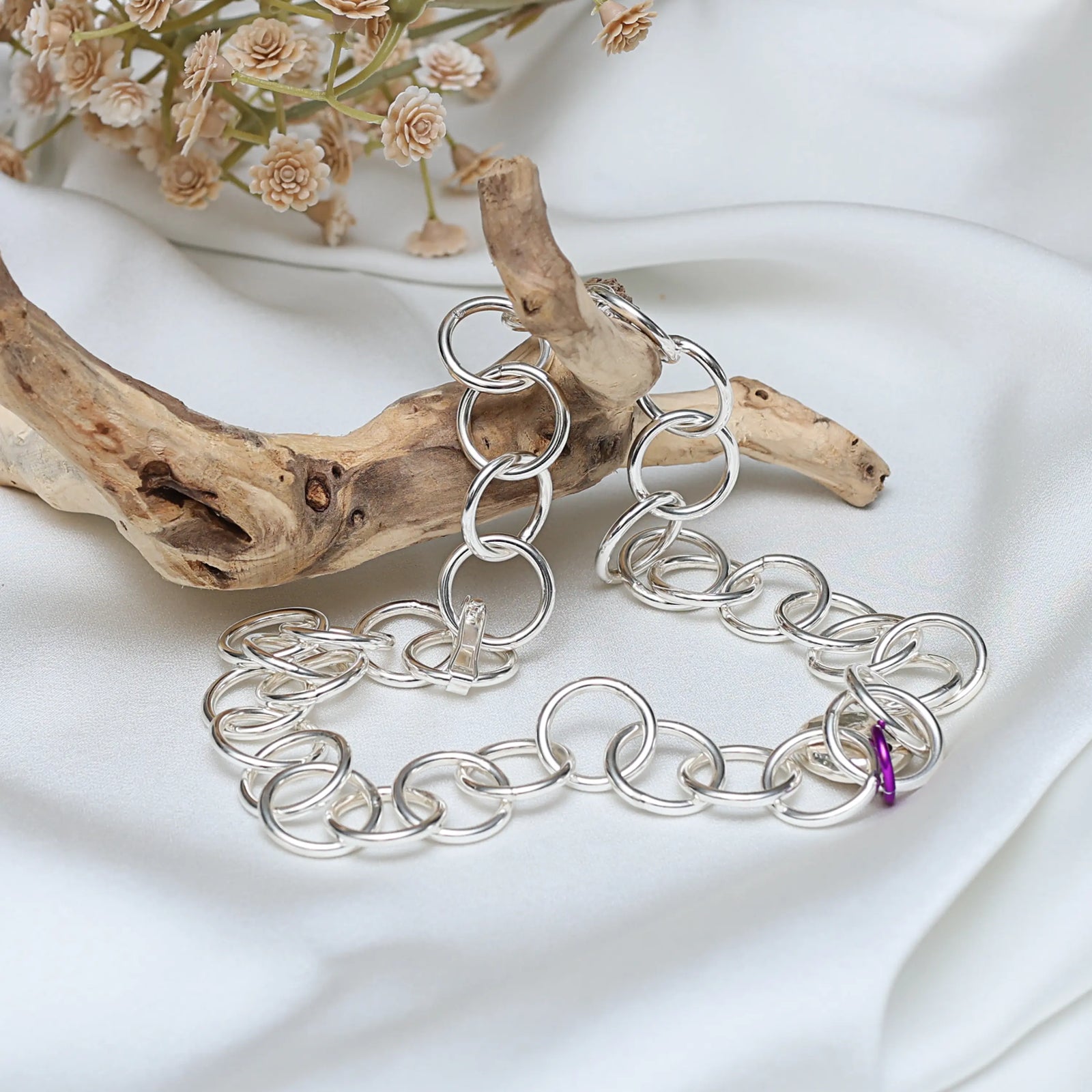 Silver chain bracelet on a white fabric background with driftwood and flowers.
